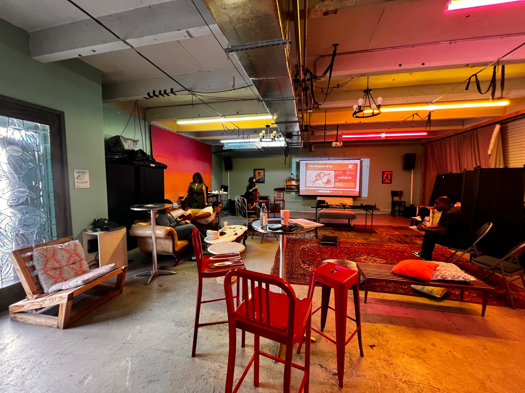 A room transformed for a HUEMUNS chamber with varied seating, art materials, warm colored light, and a presentation screen